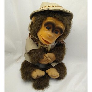 May include: A plush monkey hand puppet with dark brown fur, a tan face, and large dark eyes. The puppet wears a tan safari-style hat and a matching tan shirt with a tag that reads PUPPET. The monkey has a soft, textured appearance.