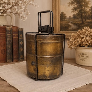 May include: A cylindrical, two-tiered brass tiffin carrier with a weathered, aged patina. The container features a dark metal frame and a central handle mechanism that secures the stacked compartments together in a vertical, upright position.