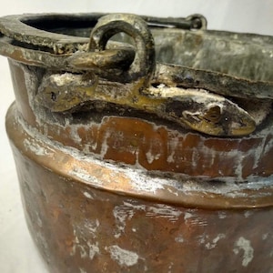 May include: A weathered copper bucket featuring a prominent metal handle attachment with a textured, oxidized surface. The vessel displays a deep brown patina with visible signs of wear, including white mineral deposits and aged metallic discoloration.