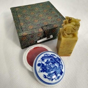 Könnte beinhalten: Ein antikes chinesisches Siegelset mit einem geschnitzten Steinsiegel mit Drachenmotiv, einem roten Stempelkissen und einer dekorativen Box. Der Deckel des Stempelkissens ist weiß mit einem blauen Drachendesign.