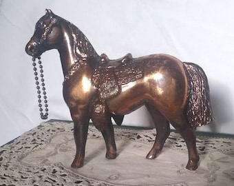 May include: A bronze-colored horse figurine with a saddle and bridle. The horse is standing on a decorative surface, and a beaded chain hangs from its mouth. The figurine has a metallic finish and detailed features.