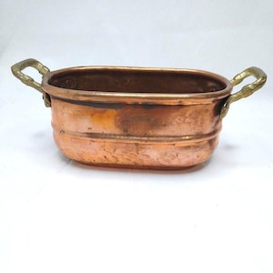 May include: An oval copper basin featuring a smooth, polished finish with visible signs of natural oxidation and wear. The vessel includes two textured, brass-toned metal handles attached to the sides, providing a contrast to the copper body.