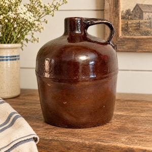 May include: A dark brown glazed ceramic jug with a rounded body, a short neck, and a single side handle. The surface features a smooth, reflective finish with subtle variations in color tone, showcasing a classic, sturdy, and functional vessel design.