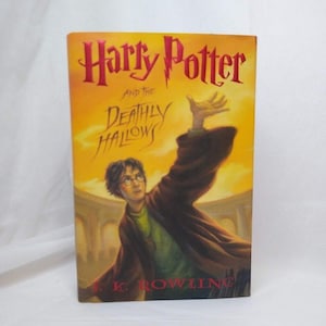 May include: A hardcover book titled Harry Potter and the Deathly Hallows by J.K. Rowling. The cover features a yellow and orange background with an illustration of a person with dark hair wearing a green shirt and brown robe reaching upward.