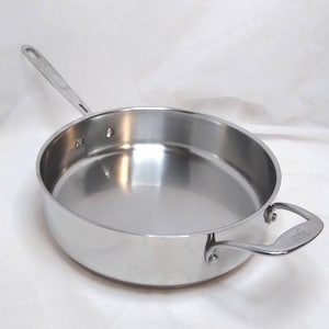 May include: A stainless steel frying pan with a long handle and a smaller loop handle. The pan has a wide, shallow cooking surface and a shiny, reflective interior. The pan is designed for cooking and is suitable for various culinary tasks.