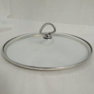 May include: A circular clear glass lid for a pot or pan, featuring a polished silver-tone metal rim and a matching silver-tone metal handle shaped like a rounded triangle, centered on the top surface of the glass lid for easy lifting.