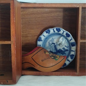 May include: A wooden display case with a Dutch theme. It contains a wooden clog with "Holland" written on it, a blue and white plate with a windmill design, and a pair of miniature white clogs. The case is made of brown wood.