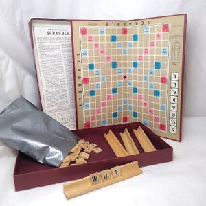 May include: Vintage Scrabble board game with a maroon box, tan game board, and wooden letter tiles. The board features blue and pink squares. Includes wooden tile holders and a rule book.