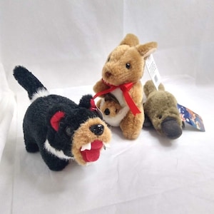 Op de afbeelding: Drie pluche dieren, waaronder een zwart-witte Tasmaanse duivel met een open bek, een bruine kangoeroe met een rood lint en een jong in de buidel, en een bruin vogelbekdier met een platte snavel, allemaal voorzien van productlabels.
