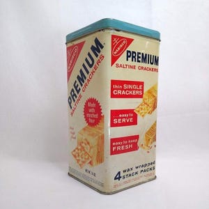 Puede incluir: Una lata de metal rectangular vintage para galletas saladas Nabisco Premium con tapa azul claro. El recipiente de color crema presenta texto en rojo y azul, incluyendo el logotipo de Nabisco, el nombre del producto y 4 wax wrapped stack packs.