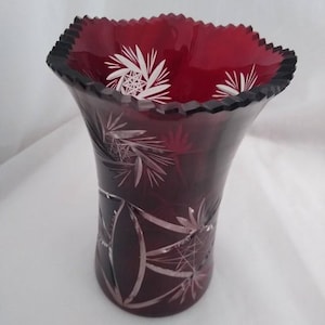 May include: A ruby red glass vase with a flared, scalloped rim. The vase features intricate white etched floral and geometric designs. The vase has a wide opening and a slightly tapered body, suitable for displaying flowers or as a decorative piece.