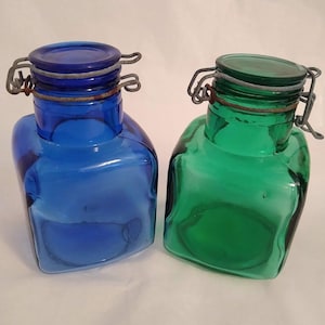 May include: Two square glass storage jars with wire bail closures. One jar is cobalt blue and the other is emerald green. Both containers feature a rounded neck, a flat glass lid, and a metal swing-top mechanism that secures the lid to the jar.