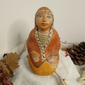 May include: A hand-painted ceramic figurine of a seated person wearing a rust-colored robe and a patterned headdress. The figure has a yellow skirt and tan skin. The figurine is on a white cloth with autumn leaves and a pinecone.