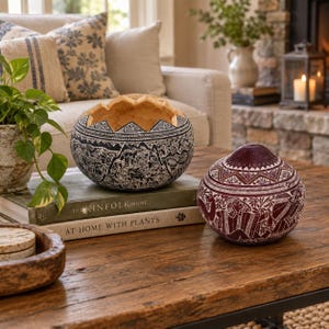 May include: A round, black and white carved gourd bowl with a jagged top edge and intricate etched patterns, paired with a smaller, round, maroon and white carved gourd container featuring a pointed lid and detailed etched designs.