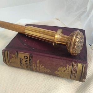 May include: A wooden walking stick with a polished brass handle featuring a fluted shaft and a round, floral-embossed top, resting on a burgundy hardcover book titled History of England with gold-embossed lettering and decorative details.