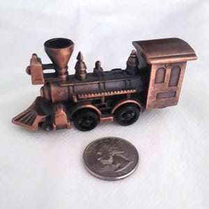 May include: A miniature copper-colored train engine with black wheels and details. The train has a smokestack, a headlight, and a small cabin. A US quarter is next to the train for size comparison.
