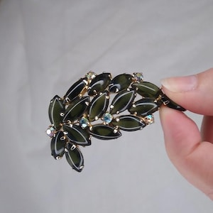 Könnte beinhalten: Vintage Brosche im Blätterdesign. Die Brosche hat dunkelgrüne, oval geformte Steine, die in einem goldfarbenen Metall gefasst sind. Kleine, schillernde Kristalle sind zwischen den grünen Steinen verstreut. Die Brosche wird von einer Hand gehalten.