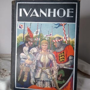 May include: A vintage hardcover book titled "Ivanhoe" with a black cover and a colorful illustration. The illustration depicts three figures in medieval armor and attire, with flags and a landscape in the background. The book is displayed on a white surface.