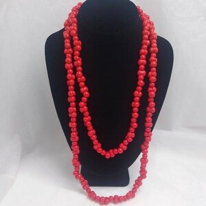 May include: A long, layered necklace composed of vibrant red beads. The beads are uniform in size and shape, strung together to create a flowing, eye-catching accessory. The necklace is displayed on a black mannequin, highlighting its color and design.