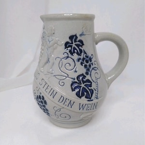May include: A stoneware pitcher with a handle, decorated with blue grape vines and leaves. The pitcher has a relief of a cherub playing a musical instrument. The text "AUS STEIN DEN WEIN" is written on a banner around the pitcher.