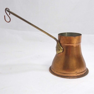 May include: A copper coffee pot with a wide, flared base and a narrow cylindrical top. It features a long, slender metal handle attached with a brass-colored plate, ending in a small, curved hook for hanging. The exterior has a warm, metallic finish.