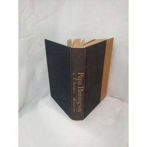 May include: A hardcover book titled "Papa Hemingway" by A.E. Hotchner. The book has a dark brown spine with gold lettering and a black cover. The pages are aged, and the book is open.