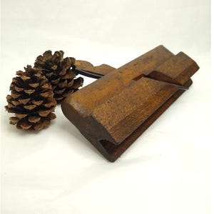 May include: A vintage wooden hand plane with a dark brown finish and a metal blade insert, positioned next to two natural brown pine cones. The tool features a rectangular body with a beveled edge and a visible slot for the cutting mechanism.