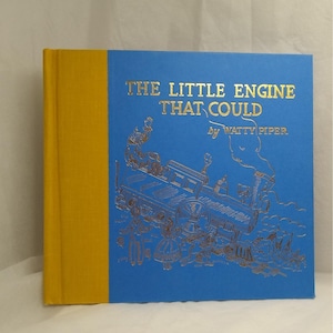 May include: A hardcover children's book titled "THE LITTLE ENGINE THAT COULD" by Watty Piper. The cover is blue with gold lettering and an illustration of a train. The spine is yellow.