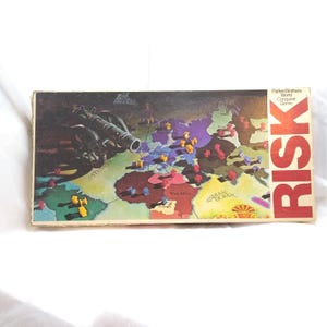 May include: A vintage Risk board game box featuring a world map with territories and game pieces. The box has a cannon illustration and the word "RISK" in large red letters. The game is by Parker Brothers.