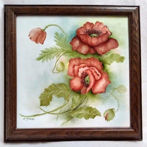 May include: A framed watercolor painting of red poppies with green leaves and buds. The artwork is set against a light blue and green background, and is enclosed in a dark brown wooden frame. The artist's signature is visible in the bottom left corner.