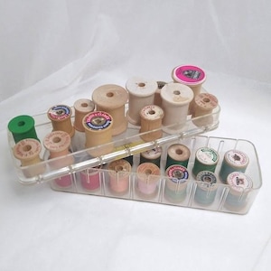 May include: A clear plastic organizer filled with vintage wooden spools of thread in various colors. The spools are arranged in two trays, showcasing a variety of thread colors and brands, perfect for sewing and crafting projects.
