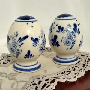 May include: Two egg-shaped salt and pepper shakers. The shakers are white with blue floral designs and blue accents on the top and base. They are sitting on a white lace doily.