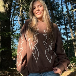 May include: A brown sweatshirt with white flame-like embroidery. The long-sleeved top is worn with blue jeans. The person is smiling in a forest setting with tall trees and sunlight.