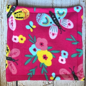 May include: A square, hot pink fleece blanket featuring a repeating pattern of butterflies and flowers. The butterflies have black bodies and wings with pink, blue, and yellow accents. The flowers are pink and yellow with green leaves.