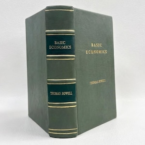 May include: A dark green hardcover book titled "Basic Economics" by Thomas Sowell. The spine and cover feature gold lettering. The spine displays the title and author's name. The book is standing upright.
