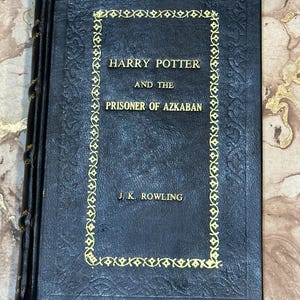 May include: A dark blue hardcover book, "Harry Potter and the Prisoner of Azkaban" by J.K. Rowling. The title and author's name are in gold lettering. The cover features a gold decorative border. The spine is black.