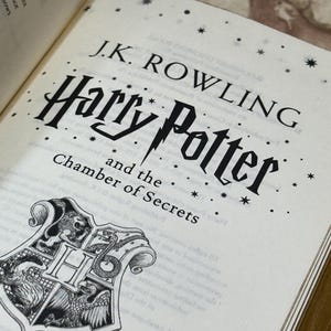 May include: An open book displaying the title "Harry Potter and the Chamber of Secrets" by J.K. Rowling. The cover features the title in a stylized font with a lightning bolt and a crest with a lion, snake, eagle, and badger.