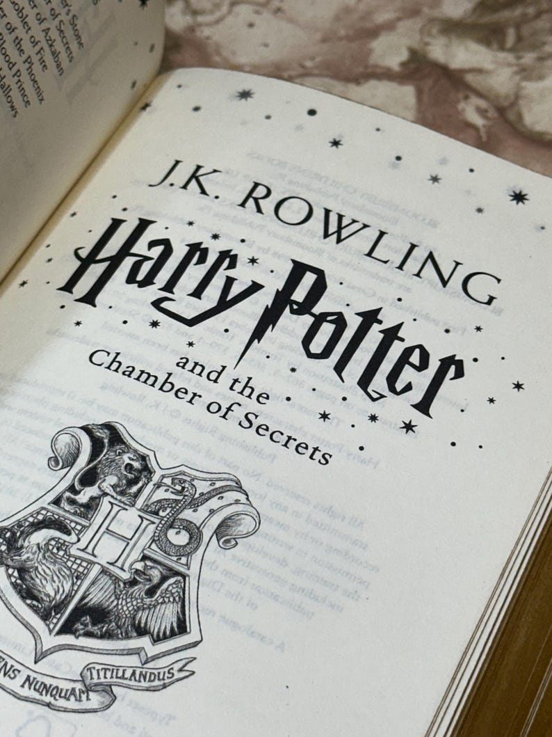 May include: An open book displaying the title "Harry Potter and the Chamber of Secrets" by J.K. Rowling. The cover features the title in a stylized font with a lightning bolt and a crest with a lion, snake, eagle, and badger.