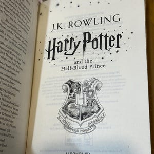 May include: An open book displaying the title "Harry Potter and the Half-Blood Prince" by J.K. Rowling. The Hogwarts crest is visible, along with the Bloomsbury Children's Books logo.