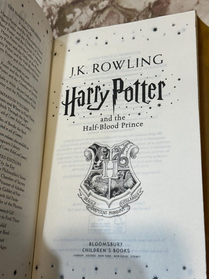 May include: An open book displaying the title "Harry Potter and the Half-Blood Prince" by J.K. Rowling. The Hogwarts crest is visible, along with the Bloomsbury Children's Books logo.