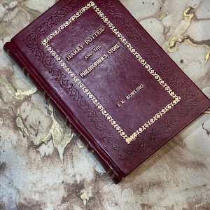 May include: A burgundy hardback book titled "Harry Potter and the Philosopher's Stone" by J.K. Rowling. The title and author's name are in gold lettering, with gold decorative trim. The book is closed and lies on a marble surface.