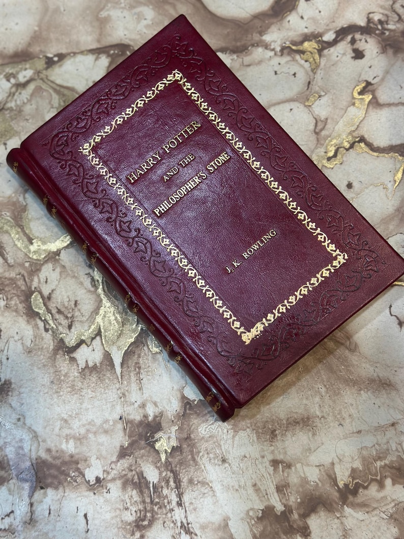May include: A burgundy hardback book titled "Harry Potter and the Philosopher's Stone" by J.K. Rowling. The title and author's name are in gold lettering, with gold decorative trim. The book is closed and lies on a marble surface.