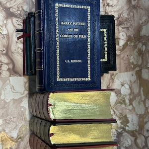 May include: A collection of books, including "Harry Potter and the Goblet of Fire" by J.K. Rowling, with gold lettering on a dark blue cover. The books have gold-gilded edges and burgundy covers. Other books are visible in the background.