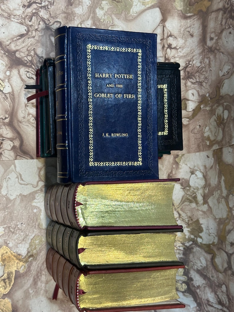 May include: A collection of books, including "Harry Potter and the Goblet of Fire" by J.K. Rowling, with gold lettering on a dark blue cover. The books have gold-gilded edges and burgundy covers. Other books are visible in the background.