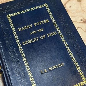 May include: A dark blue hardcover book, "Harry Potter and the Goblet of Fire" by J.K. Rowling. The title and author's name are in gold lettering. The cover features a gold decorative border and embossed detailing.