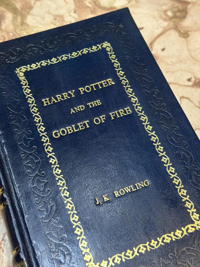 May include: A dark blue hardcover book, "Harry Potter and the Goblet of Fire" by J.K. Rowling. The title and author's name are in gold lettering. The cover features a gold decorative border and embossed detailing.