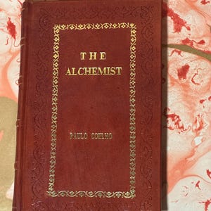 May include: A hardcover book titled "The Alchemist" by Paulo Coelho. The book has a deep red cover with gold lettering and decorative borders. The title and author's name are prominently displayed.