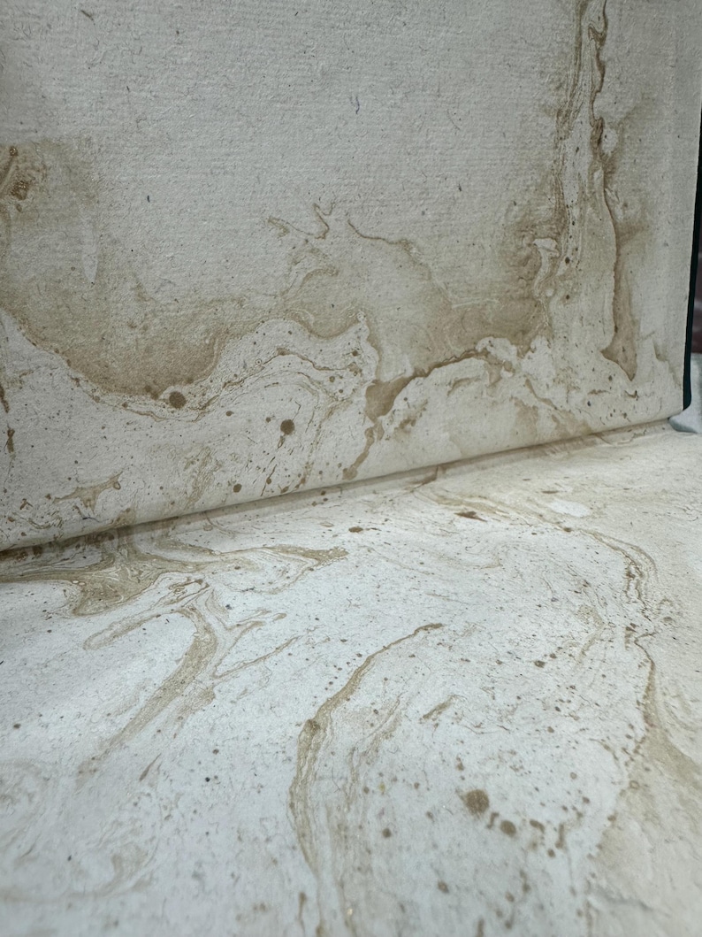 Puede incluir: Primer plano de papel texturizado blanquecino con veteado y motas marrones. El papel est&aacute; parcialmente enrollado, revelando el patr&oacute;n. El dise&ntilde;o se asemeja a la piedra natural o a un efecto acuarela, adecuado para papeler&iacute;a o proyectos art&iacute;sticos.