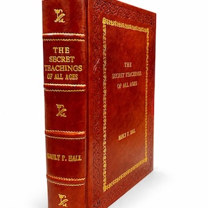 Op de afbeelding: Een groot, roestkleurig in leer gebonden boek getiteld The Secret Teachings of All Ages door Manly P. Hall. De kaft en rug zijn voorzien van goudkleurige letters en decoratieve randen, met verhoogde ribben op de rug van het boek.