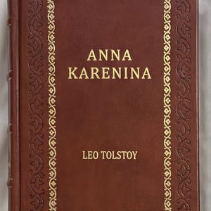 May include: A brown leather-bound book with the title "ANNA KARENINA" by Leo Tolstoy in gold lettering. The cover features a decorative gold border. A classic novel.